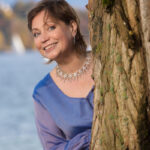 Olga Valmond im blauen Kleid an einem Baum am See – Cover der Hörprobe «O salutaris hostia» (Rossini)